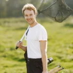 Smiling man standing outdoors holding a fishing net over his shoulder and two fishing rods in one hand, with green grass and trees in the background.
