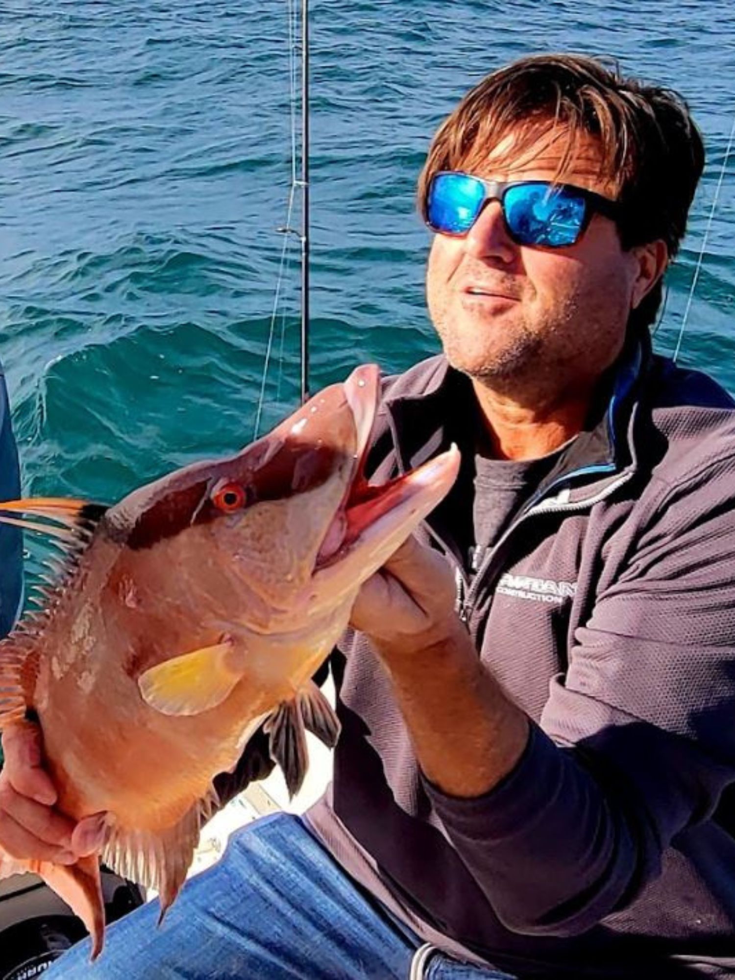 Man wearing sunglasses holds a large, colorful fish on a boat with blue water in the background.