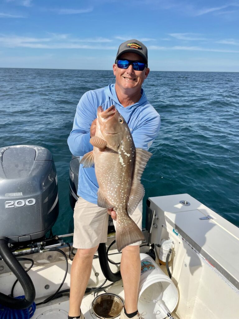 A man on a boat holding a large grouper fish with one hand, smiling at the camera, with the ocean and blue sky in the background.