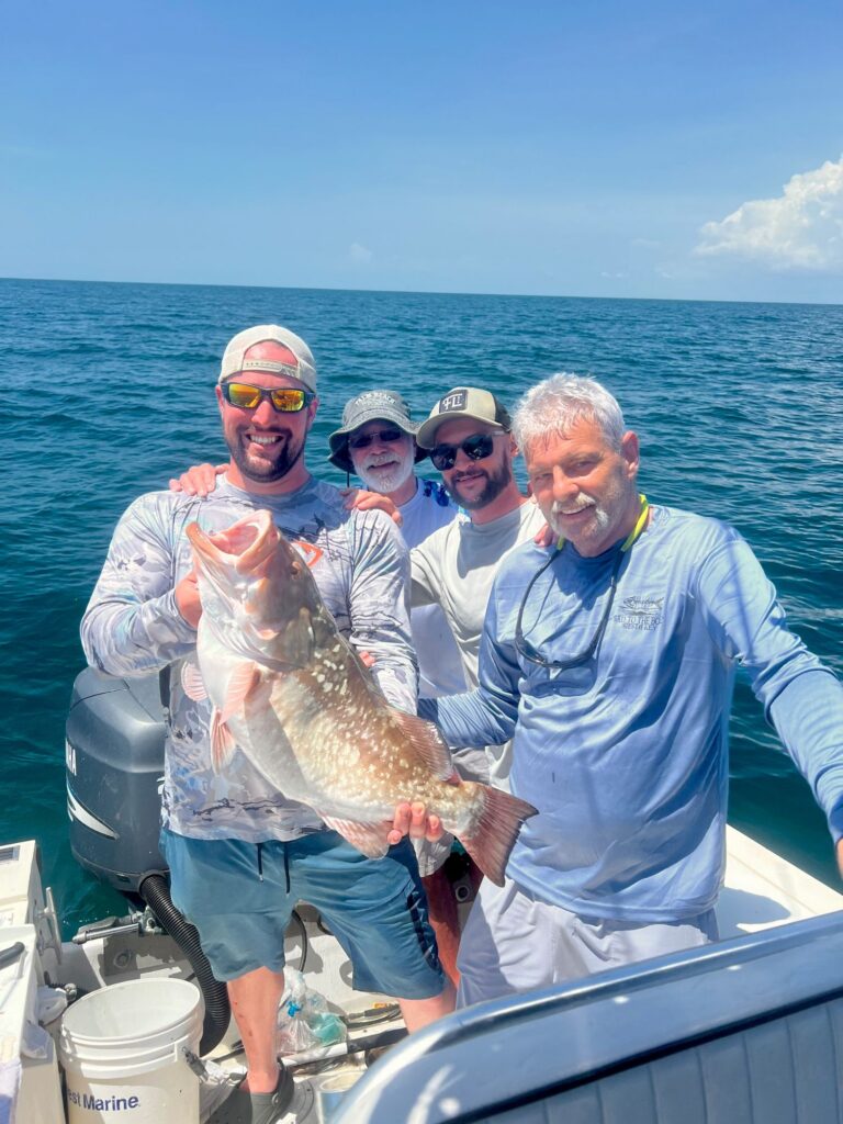 Four men on a boat pose for a photo on a sunny day, with one man holding a large fish. The ocean and sky are visible in the background.