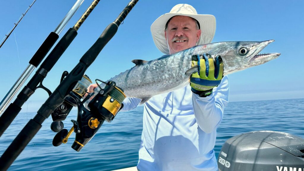 Man in a white hat and gloves holding a large fish on a boat with fishing rods, with the ocean and blue sky in the background.