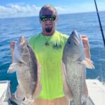 Man wearing a neon green shirt and sunglasses stands on a boat holding two large fish, with the ocean and sky in the background.