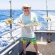 A man in a hat and striped shorts stands on a boat holding a fish, with blue ocean and fishing rods in the background.