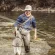 A man wearing waders and sunglasses stands in a river holding a fishing net with a fish inside.