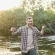A man standing in a river, smiling, holding a fishing rod in one hand and a fishing net in the other against a backdrop of trees.